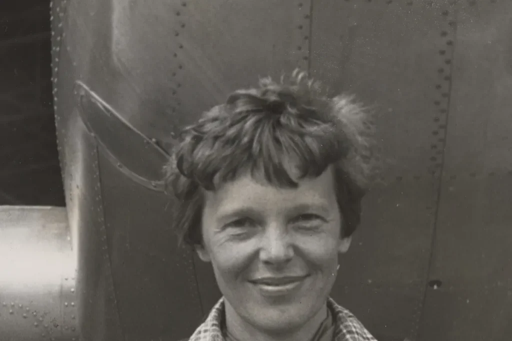 Black-and-white photograph of Amelia Earhart smiling, standing in front of the riveted metal nose of her Lockheed Electra aircraft.