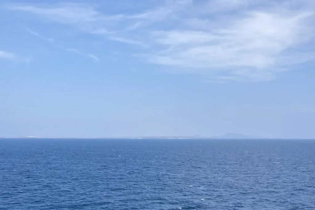 Open blue sea under a light cloudy sky with a faint low coastline on the horizon in the Bab el-Mandeb strait.