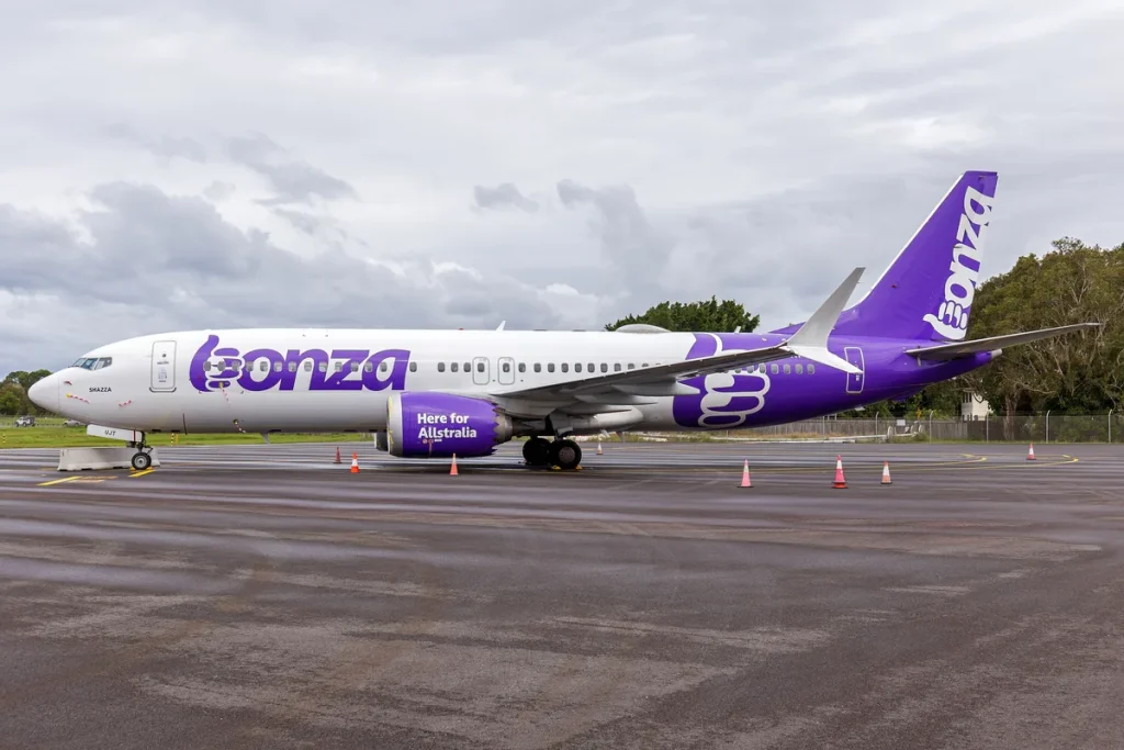 A purple and white Bonza Boeing 737 MAX 8 parked on a wet airport apron with orange safety cones, trees and a perimeter fence behind, under an overcast sky.
