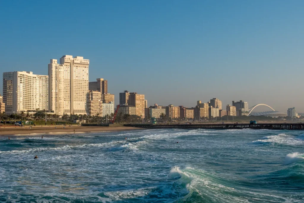 Durban's beachfront high-rises above a sandy beach and breaking surf in early morning light, with a stadium arch on the skyline at right.