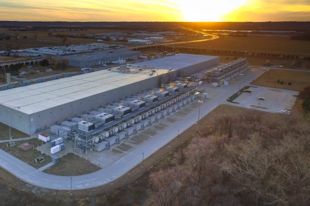 Aerial view of a long, low Google data centre building with rows of cooling units beside it, flat farmland behind, at sunset
