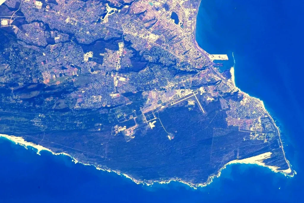 Gqeberha seen from orbit: the city spreads across the upper half of the frame, with the airport runways at centre, a dark wooded headland below and the deep blue sea of Algoa Bay curving around the coast.