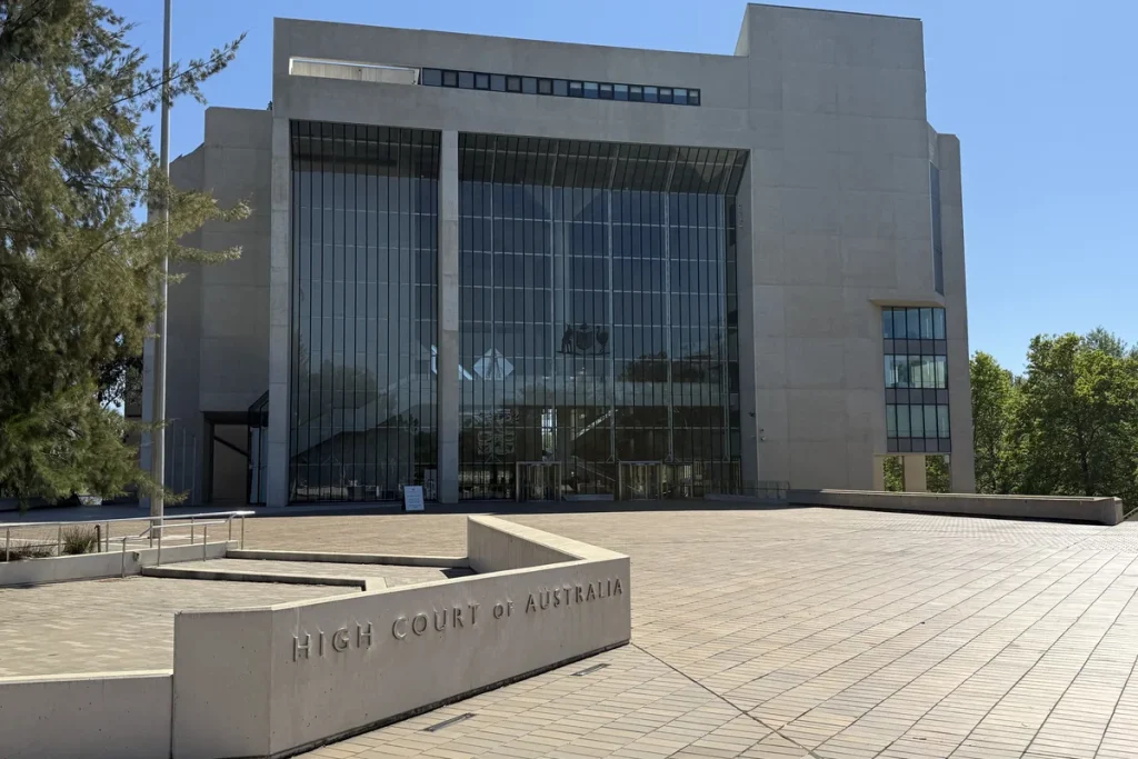 The High Court of Australia building in Canberra, its tall glass facade behind an empty paved forecourt, with the court's name carved on a low concrete wall in the foreground.