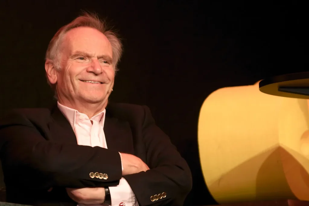 Jeffrey Archer seated on stage at the Oslo book festival in 2012, smiling with his arms folded, in a dark jacket and open-necked pale pink shirt against a black backdrop.