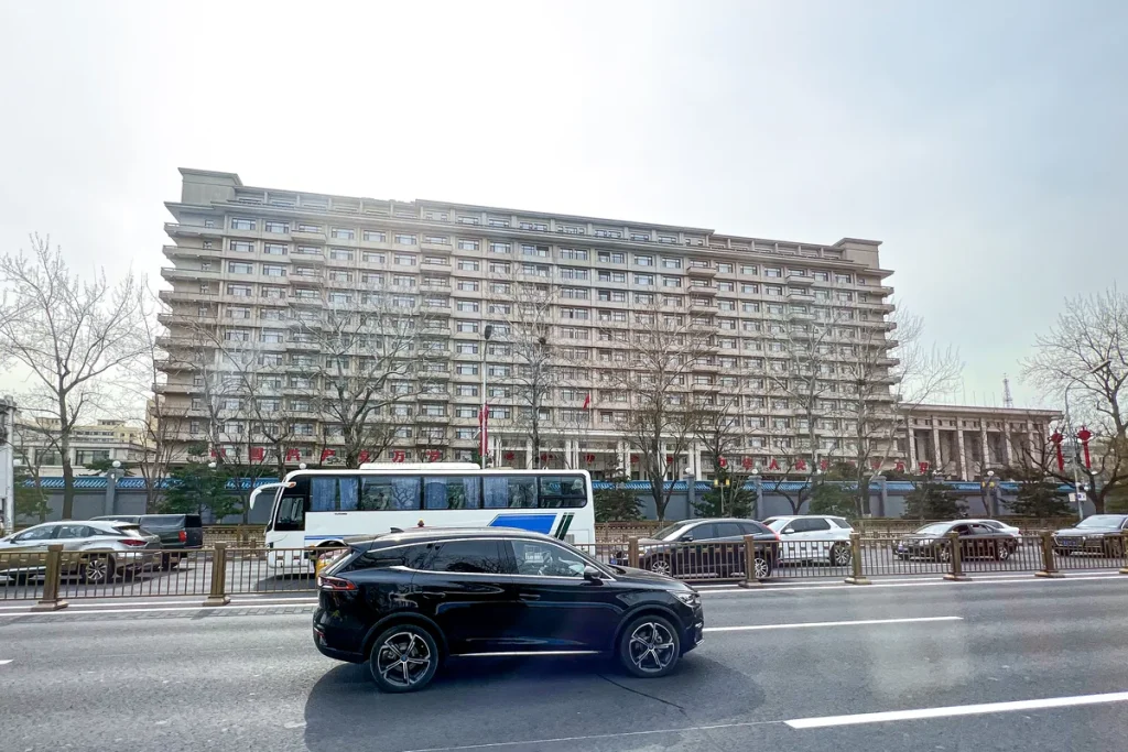 The long, multi-storey Jingxi Hotel in Beijing seen across a wide road under a pale sky, with bare trees, a white coach and a black car in front.
