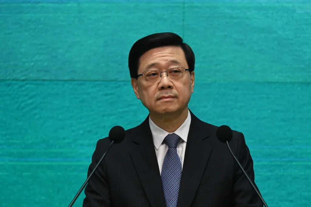 John Lee, in glasses, a dark pinstripe suit and blue patterned tie, standing behind two microphones against a teal backdrop.