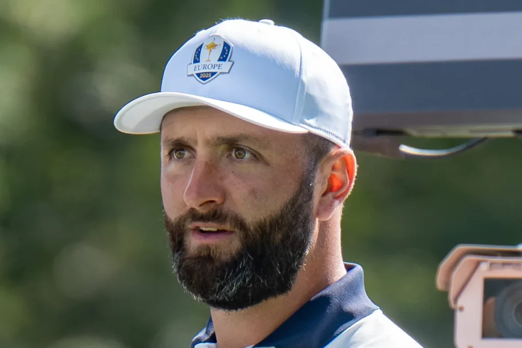 Golfer Jon Rahm in a white Europe 2025 Ryder Cup cap and a navy-collared shirt, looking off to the left, with green trees blurred behind him and part of a television camera at the right edge.
