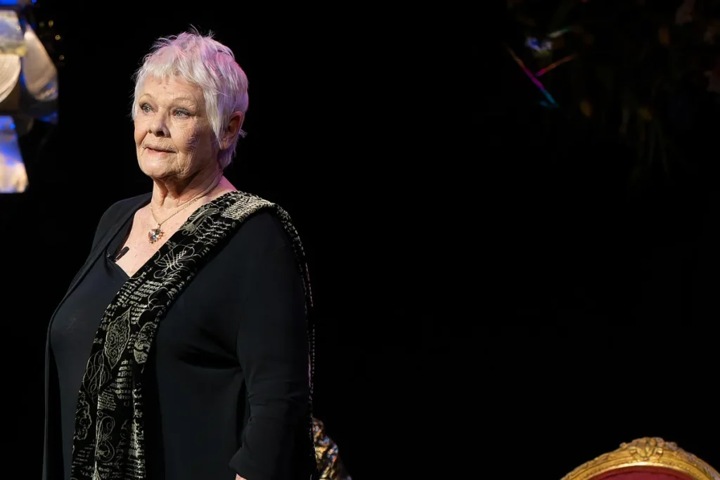Judi Dench, with short white hair, in a black top and a patterned black and gold scarf, standing on a dark stage and looking out to the audience.