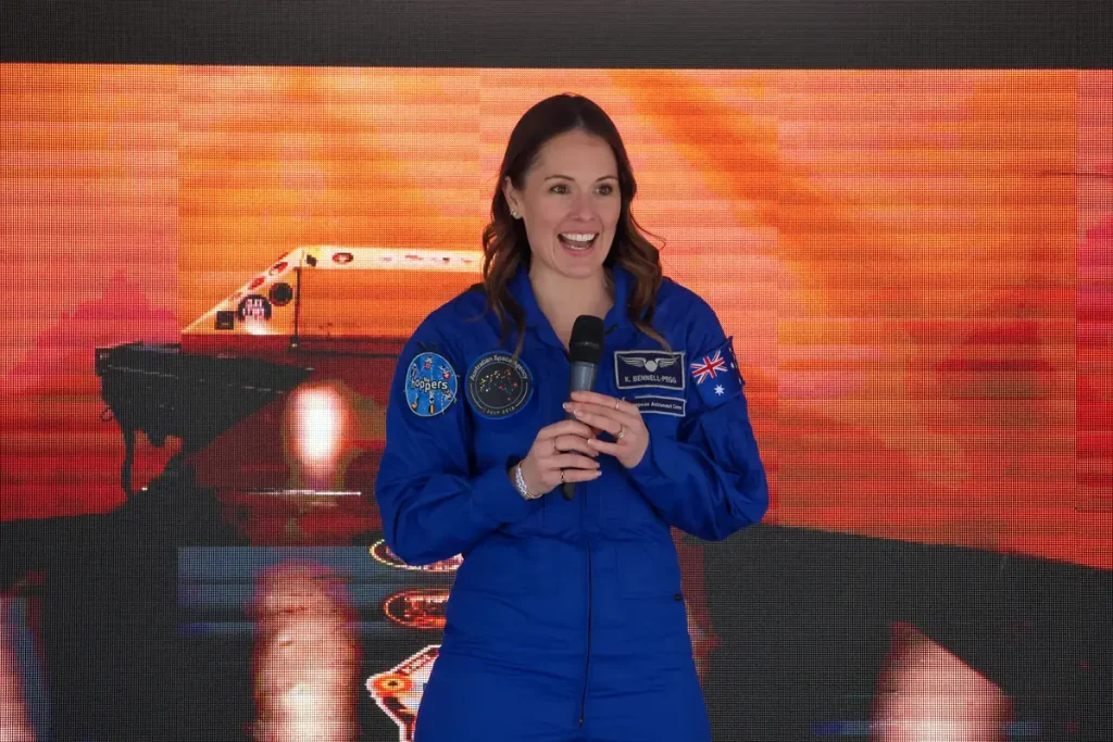 Astronaut Katherine Bennell-Pegg in a blue flight suit, holding a microphone and smiling, in front of an orange-lit stage screen.