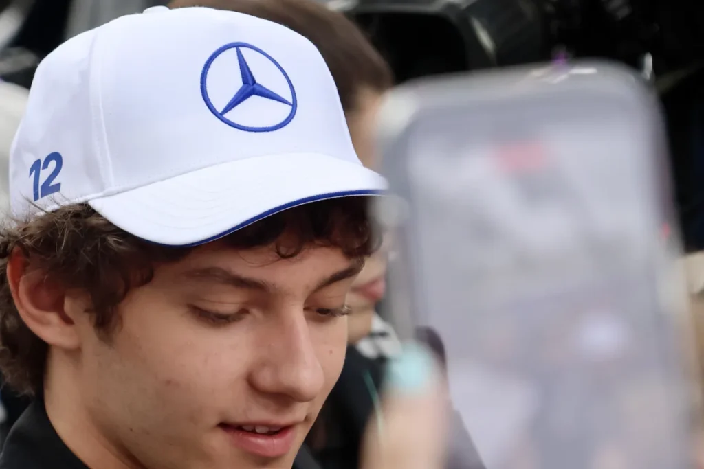 Kimi Antonelli, in a white Mercedes cap with the number 12 on the side, looks down and smiles beside a fan's raised phone.