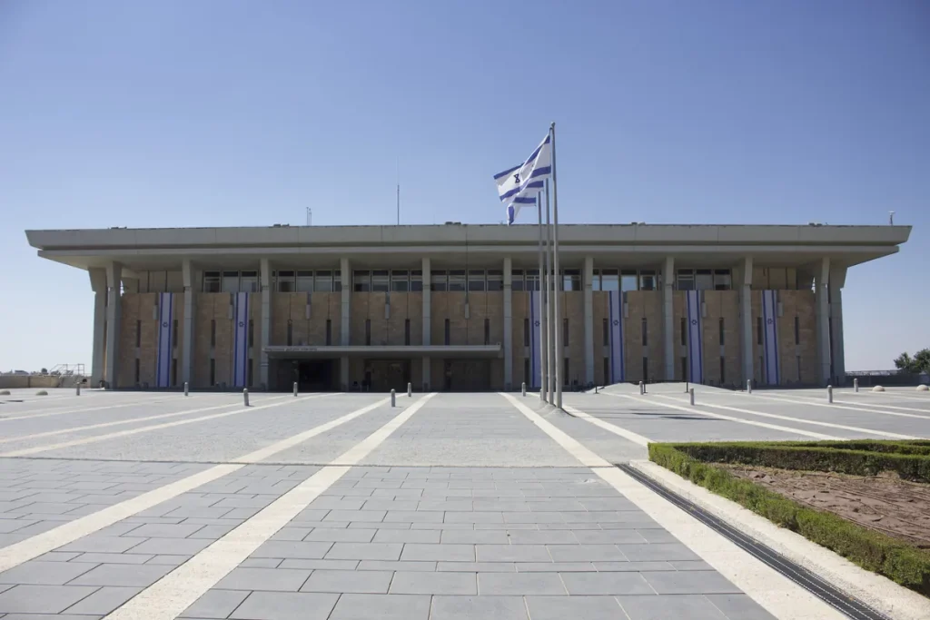 The front of the Knesset building in Jerusalem under a clear blue sky, with Israeli flags hanging between its columns and a wide paved forecourt.
