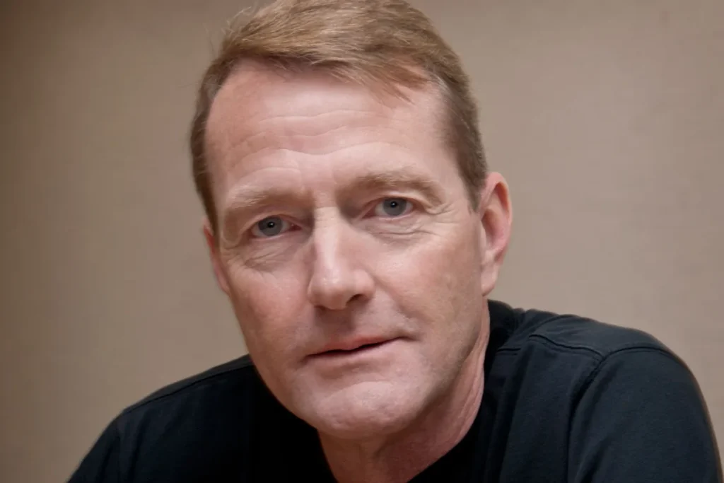 Head-and-shoulders portrait of author Lee Child, with short sandy hair and a dark crew-neck top, looking at the camera against a plain beige wall.