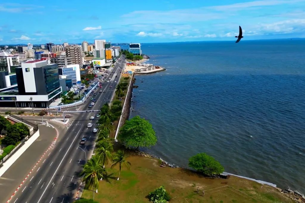 An aerial view of Libreville's seafront: a palm-lined boulevard with traffic beside a low sea wall and blue water, with office towers along the shore.