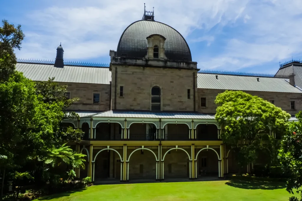 The sandstone rear wing and copper dome of Queensland's Parliament House in Brisbane, seen across a green lawn under a blue sky