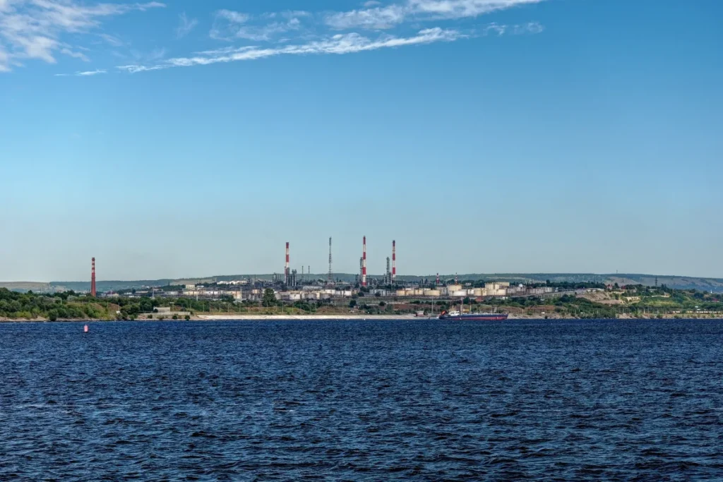 An oil refinery with red-and-white striped chimneys and pale storage tanks on the far bank of a wide river under a blue sky, with a small tanker moored at the shore.
