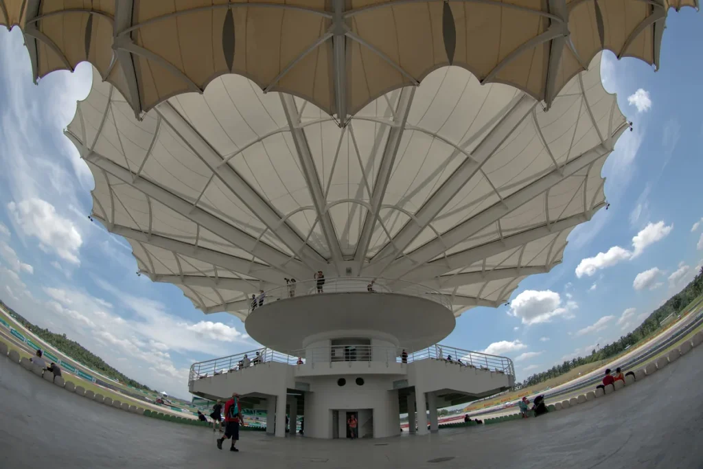A wide fisheye view beneath the white petal-shaped canopy of the grandstand tower at Sepang International Circuit, with the track curving away on both sides under a blue sky.
