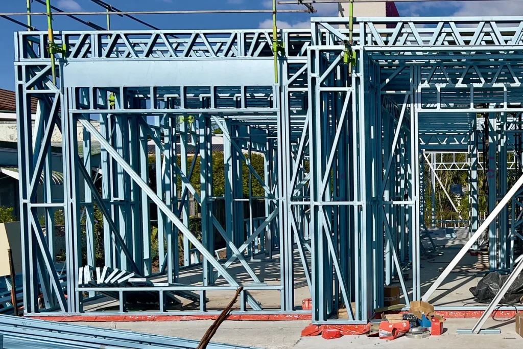 A pale blue steel house frame under construction on a concrete slab in Sherwood, Brisbane, with scaffolding along the top, roof trusses overhead, steel offcuts in the foreground and a clear blue sky.