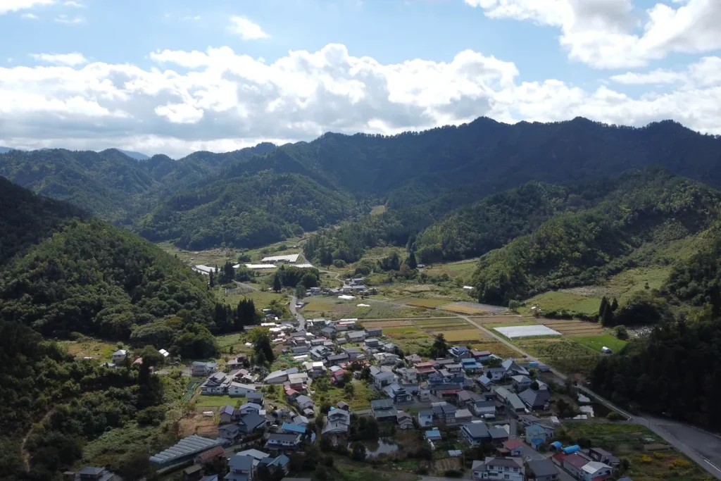 An aerial view of a village of houses and small fields in a green valley in Takahata, Japan, ringed by forested hills under white cloud.