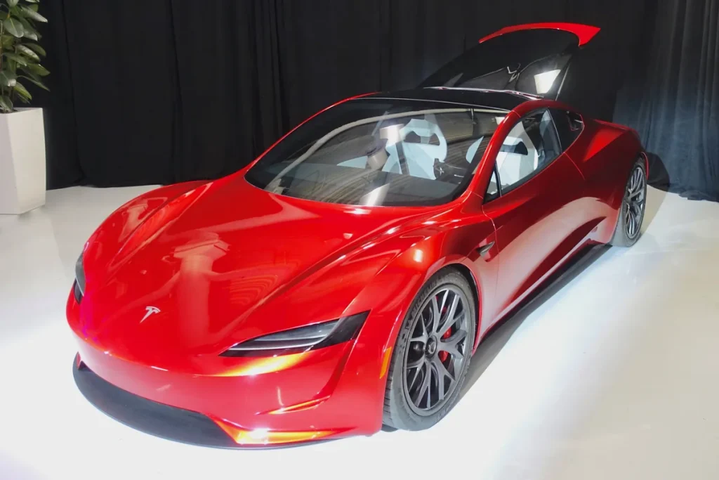 A glossy red Tesla Roadster sports car with its roof panel raised, parked alone on a white floor in front of a black curtain.