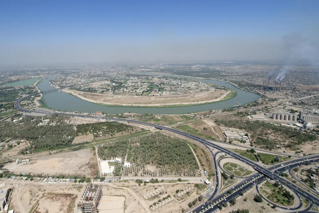 Aerial view of the Tigris River curving through Baghdad, with highway interchanges and palm groves in the foreground and power-station chimneys on the right.